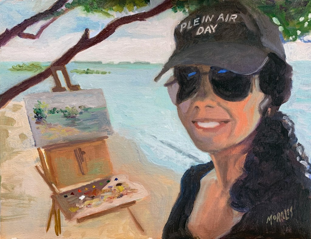 Retrato de una artista al aire libre, sonriente, con una gorra que dice 'PLEIN AIR DAY' y gafas de sol, frente a su caballete pintando un paisaje junto al agua.