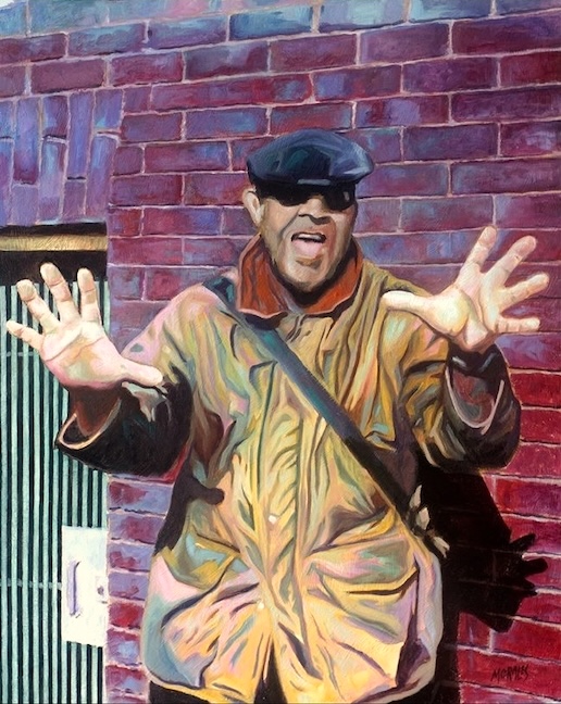 Retrato de un individuo en una chaqueta color mostaza, con un sombrero y gafas de sol, posando frente a una pared de ladrillos rojos, mostrando las manos en un gesto expresivo.